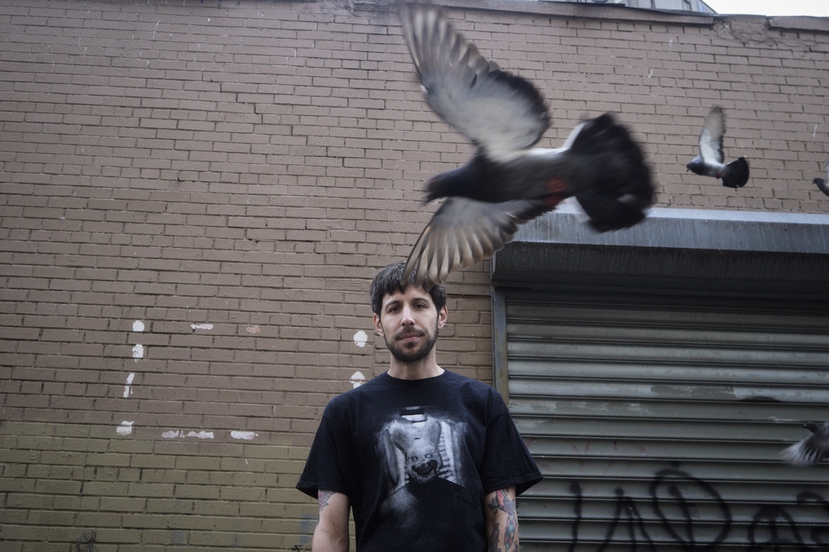 Underground hip-hop artist GDP wearing a graphic t-shirt in front of a brick wall as pigeons fly past, photographed by Rochelle Marie Adam.